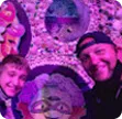 Two smiling men posing for a selfie in front of a vibrant purple floral arrangement shaped like a face.
